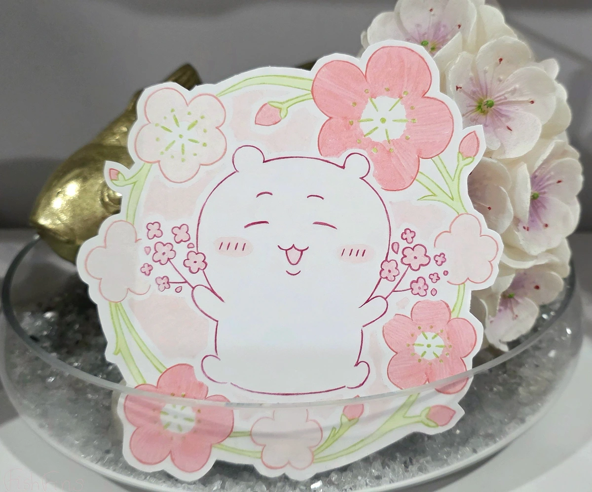 A photo of a paper birthday card coloured with alcohol markers. The card is round with a green stem outlining it and some pink flowers and flower buds of various sizes. In the middle Chiikawa is sitting and blushing with their mouth open and eyes closed. They hold onto branches with small pink flowers in both hands.